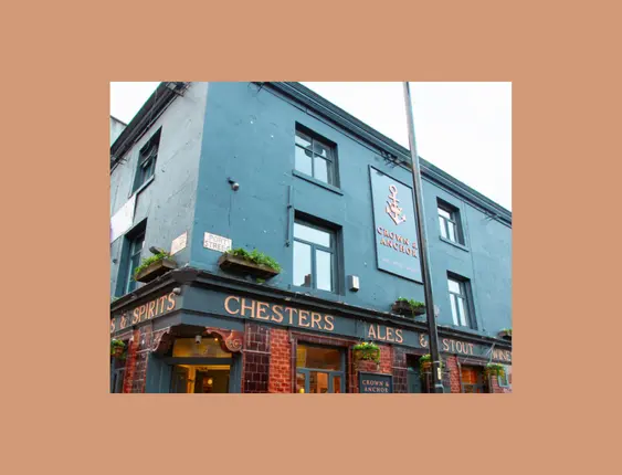 A corner pub painted blue - with a sign saying "Crown & Anchor" on the facing side. It has a banner wrapping around it, listing Ales, Stout, Wines & Spirits.