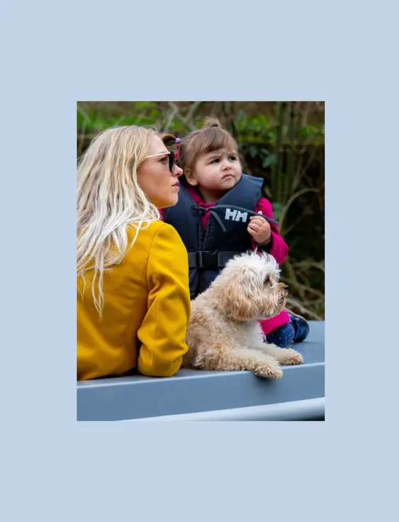 Women in yellow coat and sunglasses, child in safety jacket, and blond curly haired dog, sitting in a blue boat.
