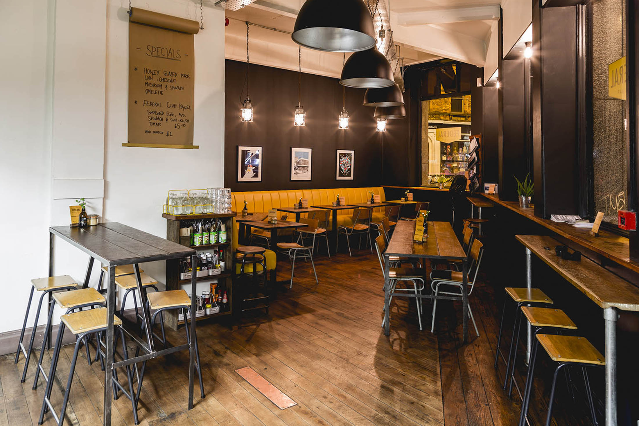A cosy café interior with dark walls, large black pendant lamps, and wooden furniture. A menu on a wall scroll lists specials.