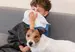A young boy sitting on a grey couch blowing his nose, with a Jack Russell dog laying in his lap