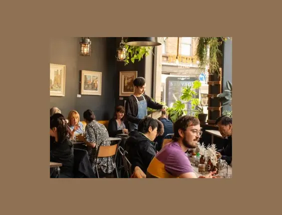 A cosy café interior with customers at various tables, decorated with hanging plants and framed pictures on dark walls, illuminated by pendant lights.