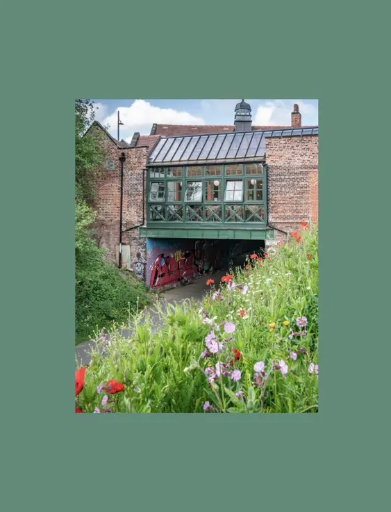 A quaint, green half-timbered building over an archway, surrounded by a vibrant wildflower foreground and lush greenery,