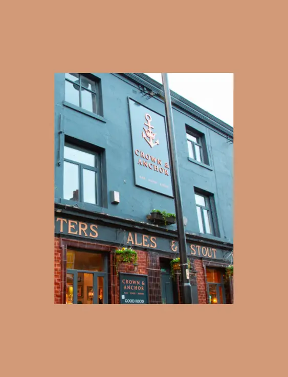 A corner pub painted blue - with a sign saying "Crown &amp; Anchor" on the facing side. It has a banner wrapping around it, listing Ales, Stout, Wines &amp; Spirits.