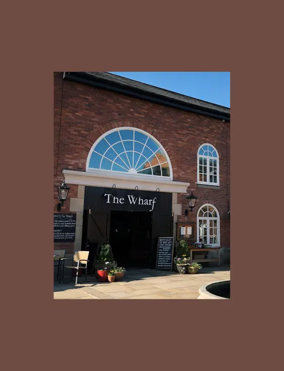 Large arched window tops the entrance of "The Wharf," a brick building with inviting outdoor seating under sunny skies.
