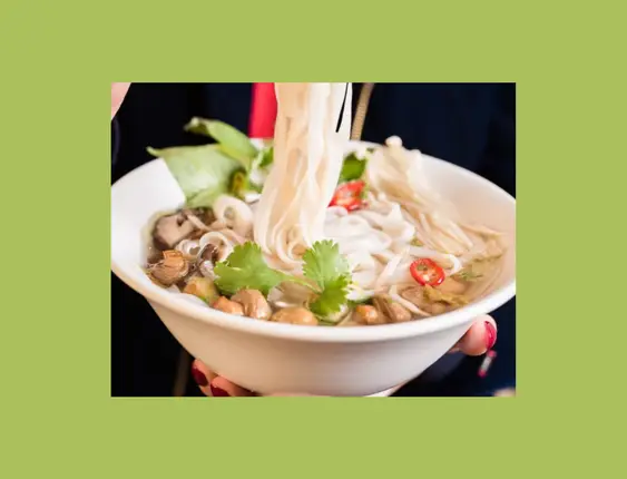 A person holds a bowl of pho, using chopsticks to lift noodles. Fresh coriander, mushrooms, and sliced chilies top this vibrant and steamy soup.