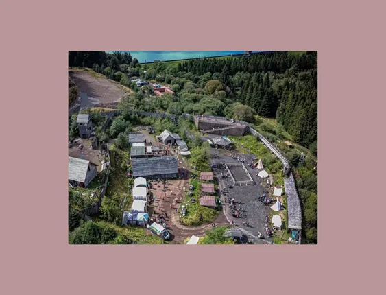 An aerial view of Duncarron Medieval Village with clustered buildings and tents, set against a backdrop of lush forests and a serene lake.