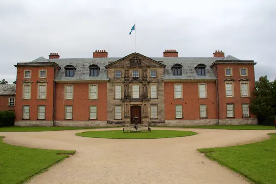 A stately Georgian manor with a symmetrical red brick façade, featuring a grand central entrance framed by ornate stonework. The manor is set against a cloudy sky, with a neatly manicured lawn and pathways leading to the entrance.