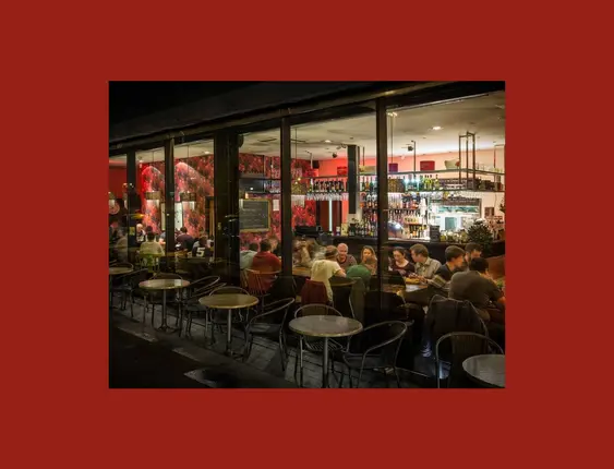Night view of a vibrant restaurant with red floral wallpaper, a bustling bar area, and patrons dining inside, seen through large glass windows.