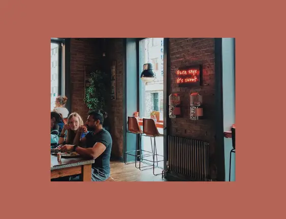 A café corner with a brick wall and a window, featuring bar stools and a neon sign saying "Ezra says it's sweet" next to hanging lamps.