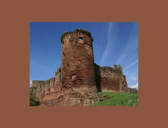 An ancient red sandstone castle with a prominent cylindrical tower stands under a blue sky, surrounded by vibrant green grass.