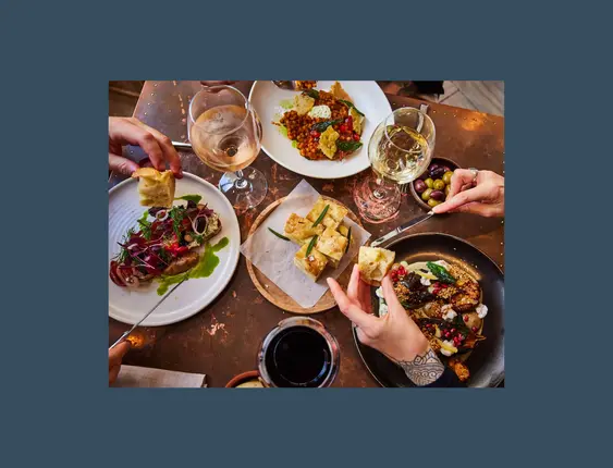 Hands reaching into table of food and wine.