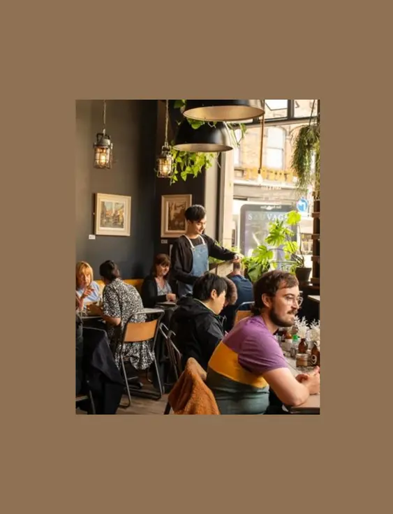 A cosy café interior with customers at various tables, decorated with hanging plants and framed pictures on dark walls, illuminated by pendant lights.