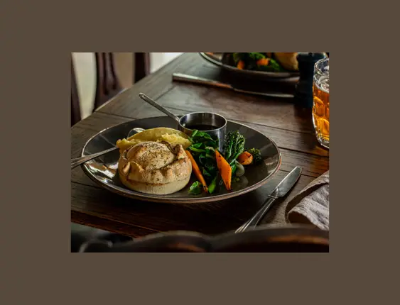 Pie, mash, vegetables and gravy on a plate, on a dark wood table.