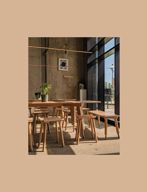 Inside a café with concrete walls, large glass windows, wooden tables, and stools; patrons sit near the entrance, enjoying the sunny view outside.