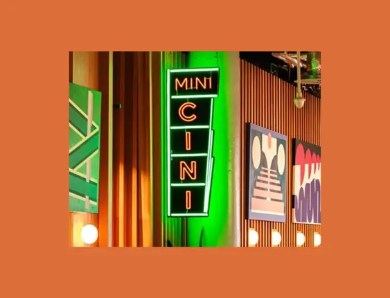 A neon sign reading "MINI CINI" in vertical orientation, mounted on a ridged green wall, illuminated in green and white light, next to wall art and under ceiling lights.