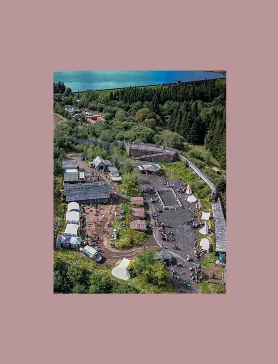 An aerial view of Duncarron Medieval Village with clustered buildings and tents, set against a backdrop of lush forests and a serene lake.