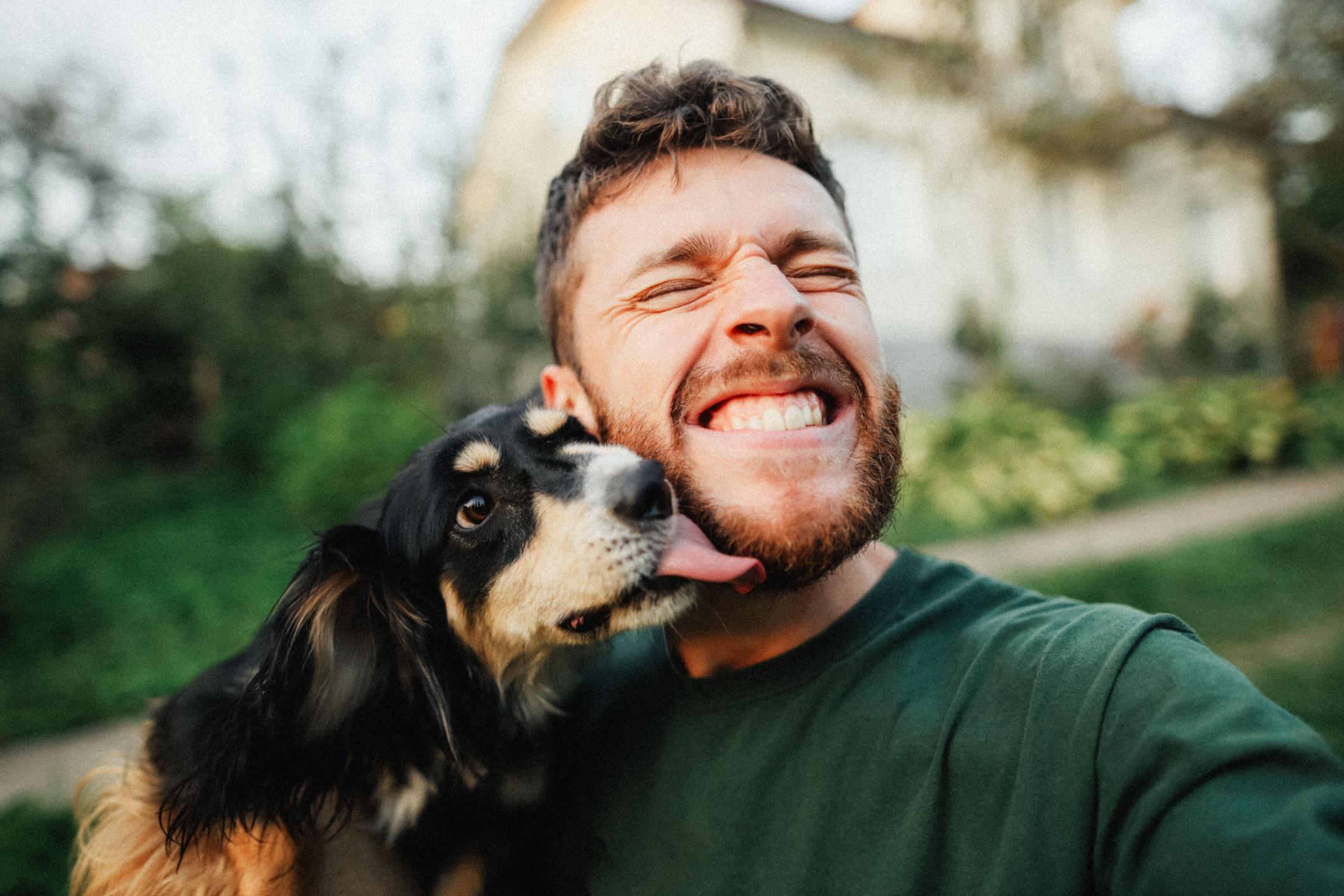 Man with a beard smiling as a dog licks his face, standing outdoors in a garden with a blurred house in the background.