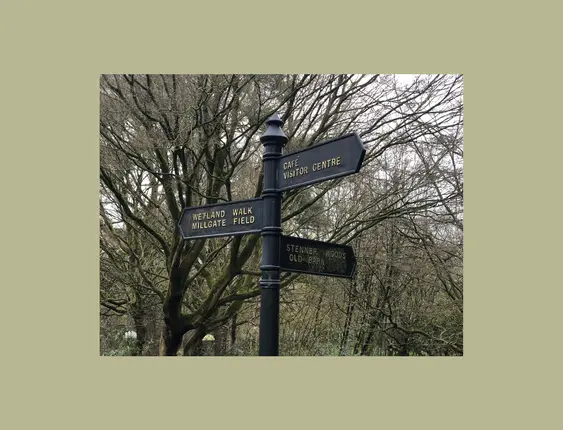 A directional signpost with arrows pointing to various locations, including "Wetland Walk", "Millgate Field","Cafe", "Visitor Centre", "Stenner Woods", and "Old Barn", set against a backdrop of leafless trees.