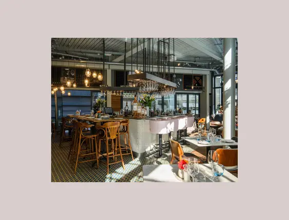 Upscale wine bar interior with circular central bar, topped with a chandelier of wine glasses. Pink upholstered bar stools line the bar, complementing the dark wooden floors and reflective mirrors that enhance the spacious feel. Ambient pendant lights cast a warm glow over neatly set dining tables