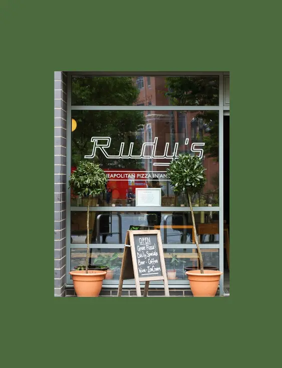 Rudy's Pizza shopfront: modern facade with large windows, featuring neon signage, open door, and street seating. A chalkboard menu stands by the entrance.