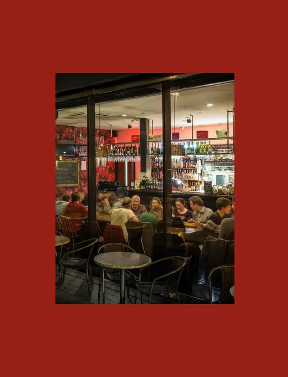 Night view of a vibrant restaurant with red floral wallpaper, a bustling bar area, and patrons dining inside, seen through large glass windows.