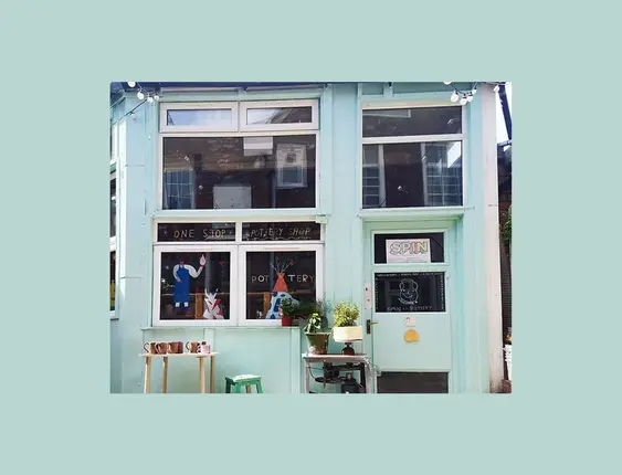 Spin Pottery Shop's pale blue facade features large windows displaying pottery and creative signs, inviting a warm, artistic vibe.