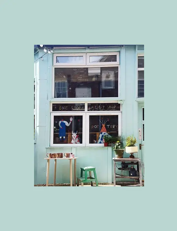 Spin Pottery Shop's pale blue facade features large windows displaying pottery and creative signs, inviting a warm, artistic vibe.