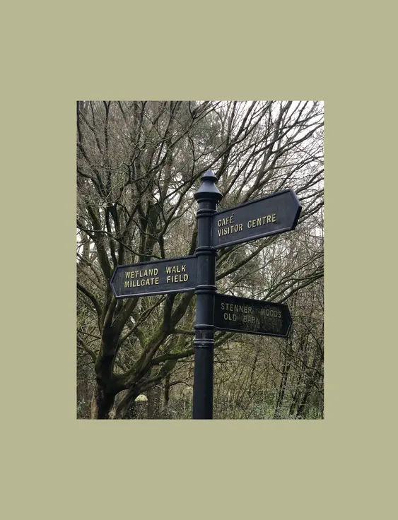 A directional signpost with arrows pointing to various locations, including "Wetland Walk", "Millgate Field","Cafe", "Visitor Centre", "Stenner Woods", and "Old Barn", set against a backdrop of leafless trees.