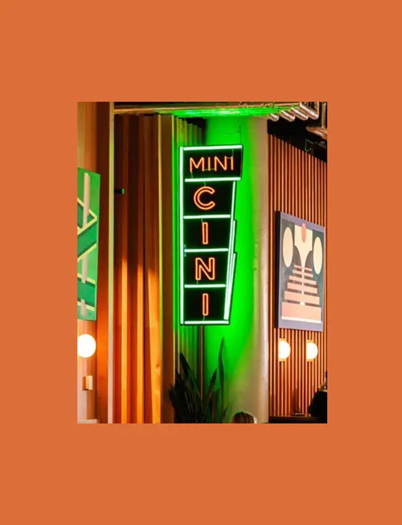 A neon sign reading "MINI CINI" in vertical orientation, mounted on a ridged green wall, illuminated in green and white light, next to wall art and under ceiling lights.