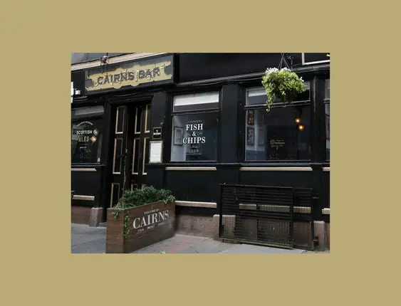 Exterior view of Cairns Bar, featuring a black facade with gold lettering and Scottish ale and fish + chip offerings.
