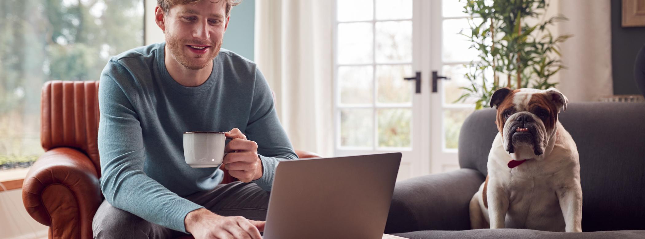 A man sits with a cup of tea next to his Bulldog while working from home