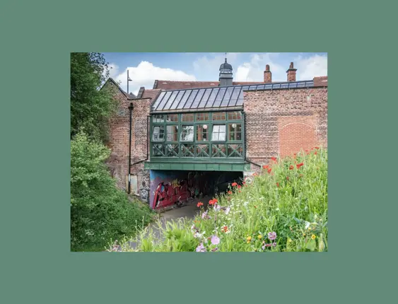 A quaint, green half-timbered building over an archway, surrounded by a vibrant wildflower foreground and lush greenery,