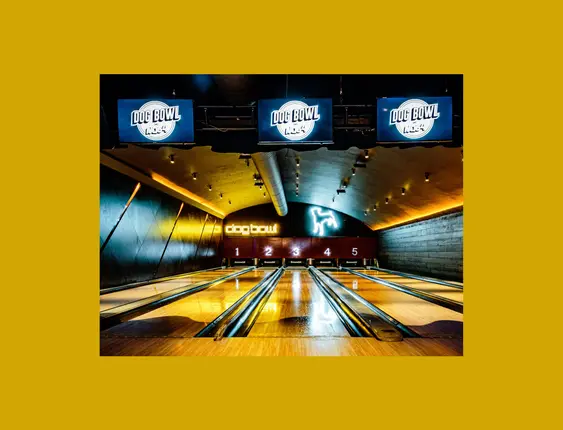 A modern bowling alley featuring five lanes under warm, ambient lighting with screens saying 'Dog Bowl' prominently displayed above each lane.