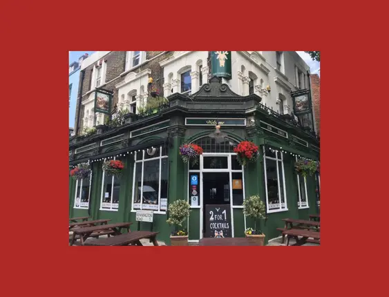 Traditional British pub on a corner, with vibrant flower baskets and '2 for 1 Cocktails' offer. The green facade stands out under a clear sky, inviting guests to its outdoor seating area.