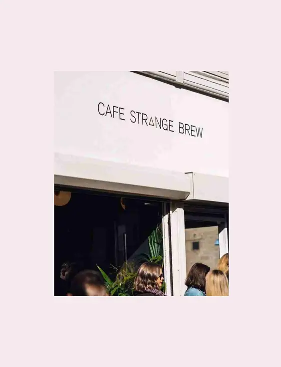 Outside Cafe Strange Brew, people wait in line under a sign. Sunlit, casual scene with plants at the cafe entrance.