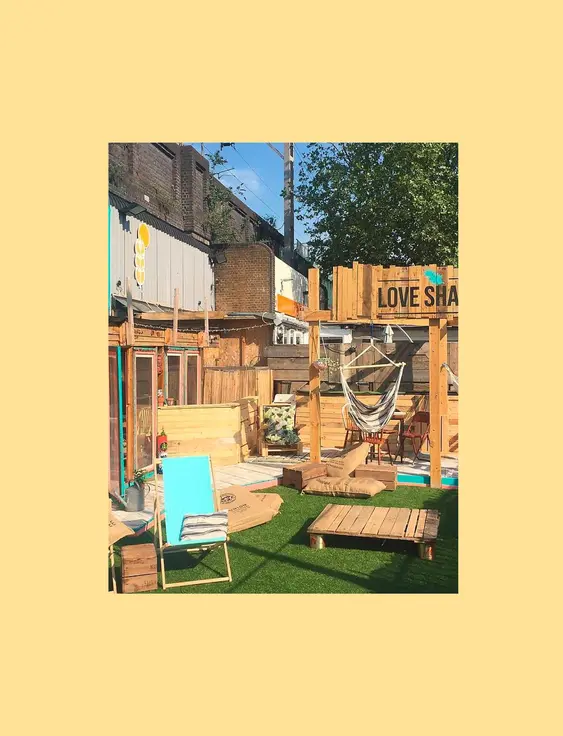 An outdoor restaurant space that shows multiple wooden decks and chairs for sitting. There is a small patch of astro turf at the front, upon which sits a blue deckchair.