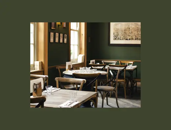 Intimate pub setting with dark green walls and wooden furniture. Natural light highlights the classic bentwood chairs and rustic tables set for dining. The walls are adorned with framed pictures and a large vintage-style painting.