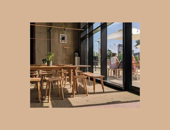 Inside a café with concrete walls, large glass windows, wooden tables, and stools; patrons sit near the entrance, enjoying the sunny view outside.