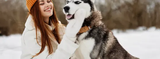 A woman with long red hair wearing an orange hat pets a husky dog outside in the snow