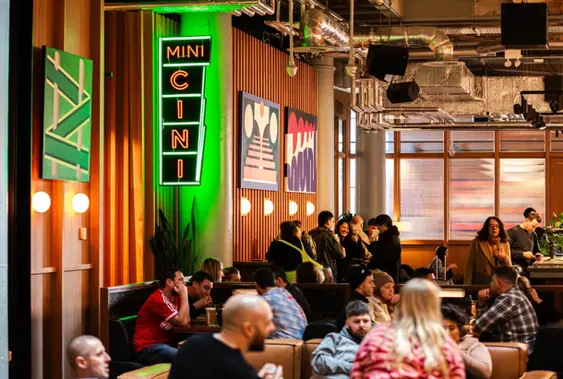 "MINI CINI" in vertical orientation, mounted on a ridged green wall, illuminated in green and white light, next to wall art and under ceiling lights.