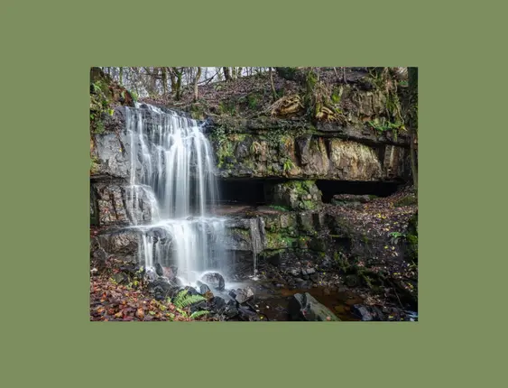 A cascading waterfall flows gracefully over mossy, rock-laden cliffs surrounded by autumn leaves and verdant foliage