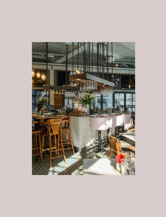 Upscale wine bar interior with circular central bar, topped with a chandelier of wine glasses. Pink upholstered bar stools line the bar, complementing the dark wooden floors and reflective mirrors that enhance the spacious feel. Ambient pendant lights cast a warm glow over neatly set dining tables