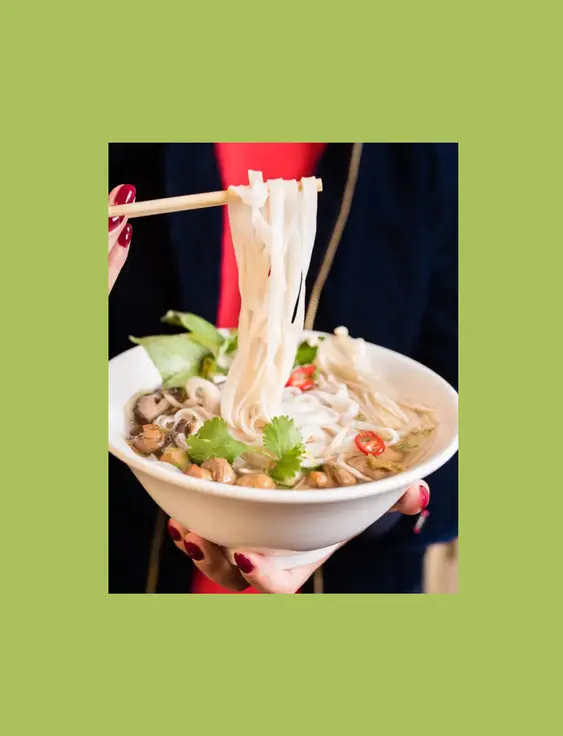 A person holds a bowl of pho, using chopsticks to lift noodles. Fresh coriander, mushrooms, and sliced chilies top this vibrant and steamy soup.
