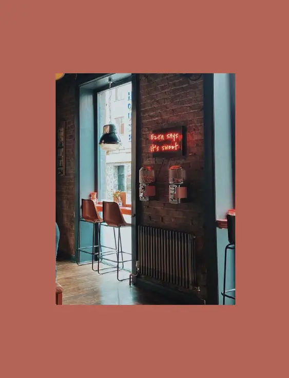 A café corner with a brick wall and a window, featuring bar stools and a neon sign saying "Ezra says it's sweet" next to hanging lamps.