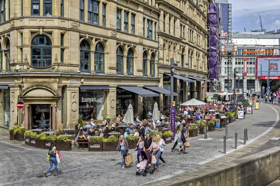 Busy urban plaza with people dining at outdoor terraces of bistros under large umbrellas, surrounded by historical buildings in the vibrant city setting of Manchester.