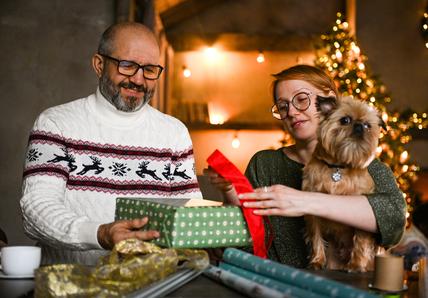 A man and a woman, likely a couple, wrap gifts while a dog is sat on the woman's lap
