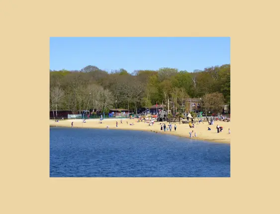Scenic view of Ruislip Lido's sandy beach and playground bustling with families enjoying a sunny day by the water, surrounded by a leafy forest