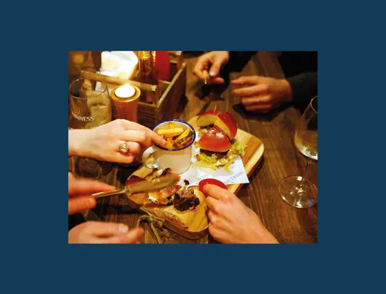 Four people's hands reaching towards a wooden board with a burger and chips on top of it.