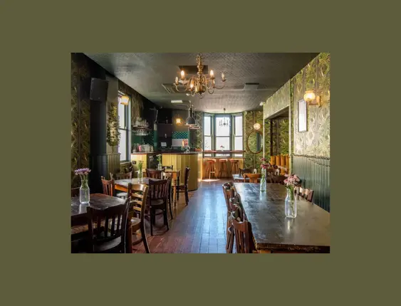 Inside of a bar with green patterned wallpaper, wooden tables and chairs.