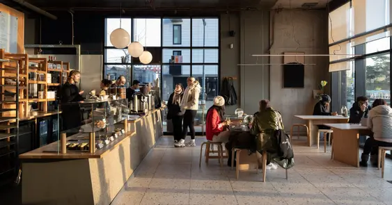Indoor coffee shop scene with large windows casting light on patrons at tables and a counter displaying pastries. A woman orders coffee while others converse or use their phones.
